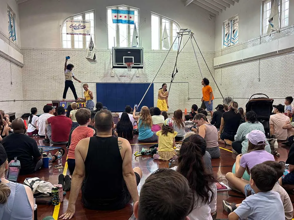 Children and adults sit on the floor of the fieldhouse gym watching circus performers, one balancing on a box and others beside an aerial rig.