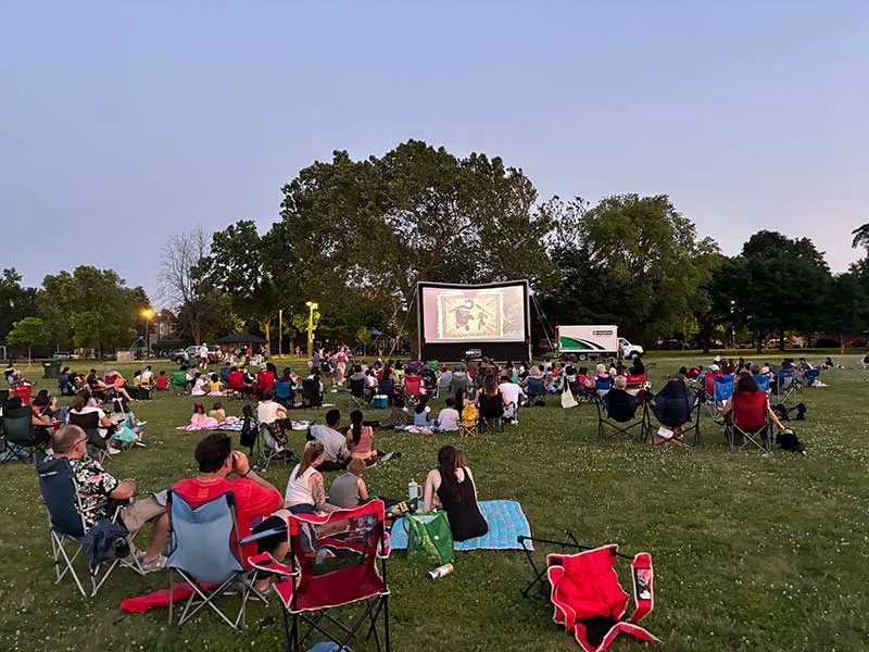 An outdoor movie in the park at dusk: a crowd on lawn chairs and blankets watches a large inflatable screen in front of tall trees.