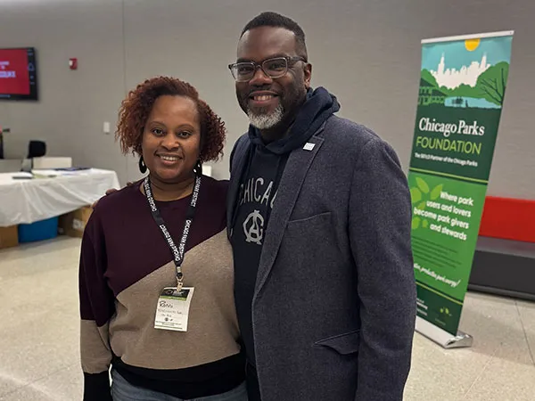 A woman wearing a conference badge stands with a smiling man in glasses and a gray blazer in front of a banner reading Chicago Parks Foundation, Where park users and lovers become park givers and stewards.