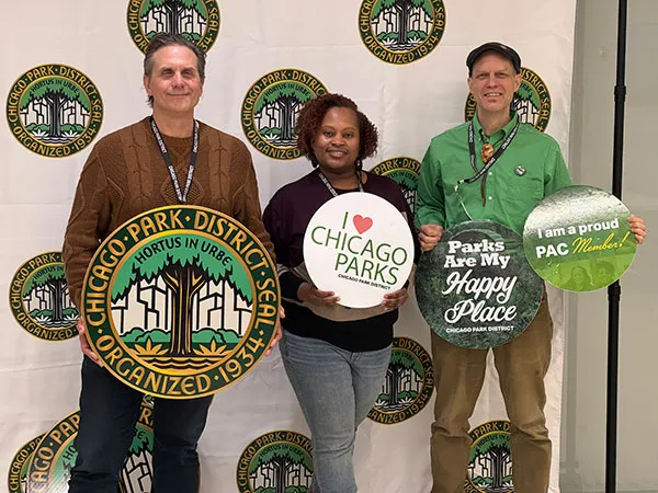 Three people pose in front of a backdrop printed with the Chicago Park District seal. One holds a large seal reading Chicago Park District Seal, Hortus in Urbe, Organized 1934; the others hold round signs reading I love Chicago Parks, Parks Are My Happy Place, and I am a proud PAC Member!