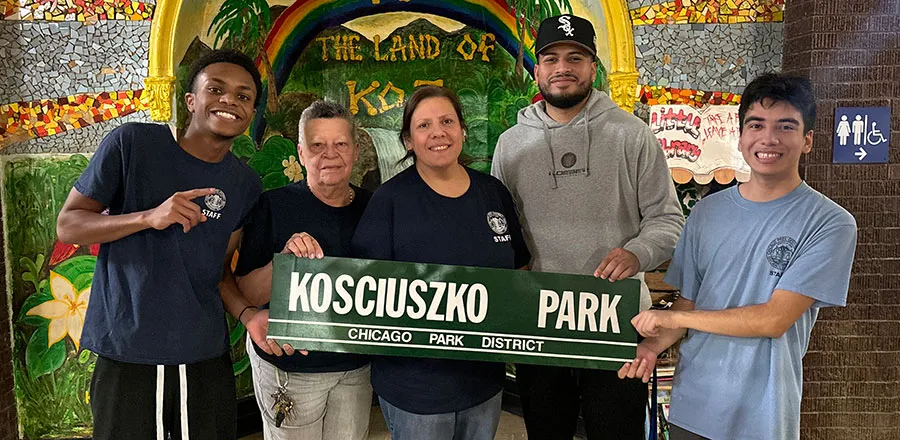 Five people, four in park staff T-shirts, hold a green sign reading Kosciuszko Park, Chicago Park District, in front of a mosaic mural with a rainbow and the words The Land of Koz.